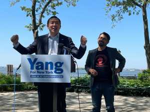 Andrew Yang with outstretched arms at a podium next to City Councilmember Carlos Menchaca in Battery Park with a view of the Statue of Liberty.