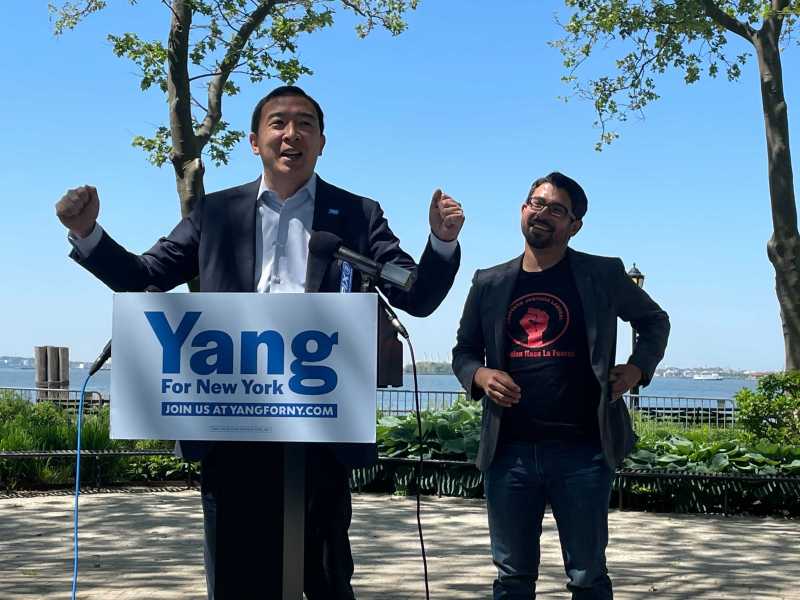 Andrew Yang with outstretched arms at a podium next to City Councilmember Carlos Menchaca in Battery Park with a view of the Statue of Liberty.