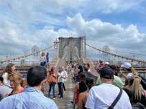 A large crowd of tourists stands on the Brooklyn Bridge walkway under sunny blue skies in July.